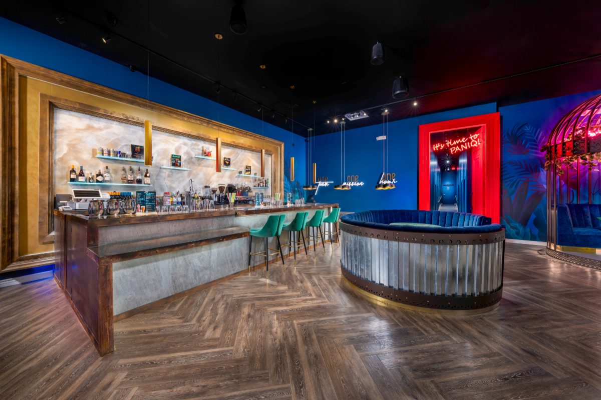 Interior of PanIQ Escape Room bar and lounge at The Venetian Las Vegas, featuring deep blue walls, a gold-framed onyx back bar with copper and concrete bar top, teal velvet bar sto