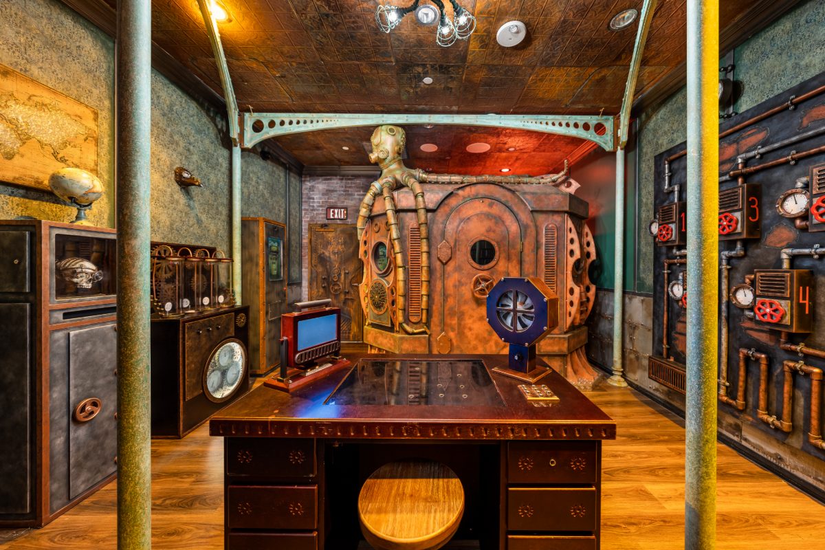 Interior of The Time Machine escape room at PanIQ Las Vegas, styled as Professor Hoffman's steampunk laboratory — a sloped pressed-bronze tin ceiling, verdigris painted columns and